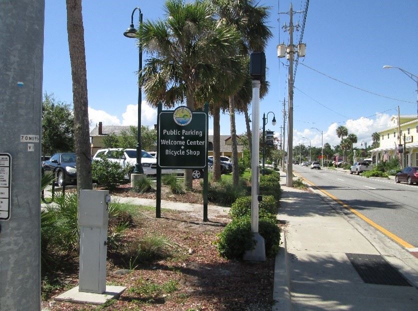 Photo of a large green sign in front of a parking lot, 

which reads: Public Parking - Welcome Center - Bicycle Shop