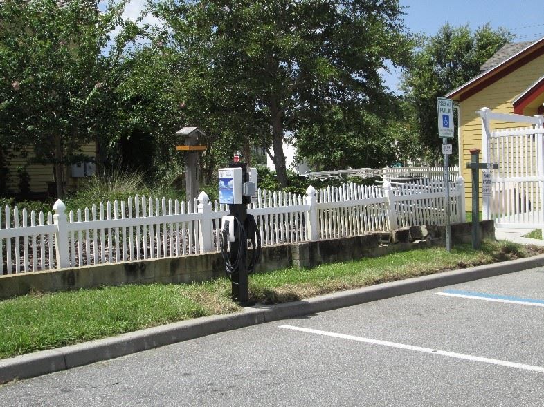 Photo of an electric vehicle charging station next to a white fence, in a parking lot.