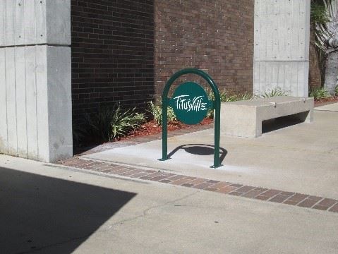 Photo of a green rounded bike rack with the name Titusville in stylized font, in front of a brick 

building.