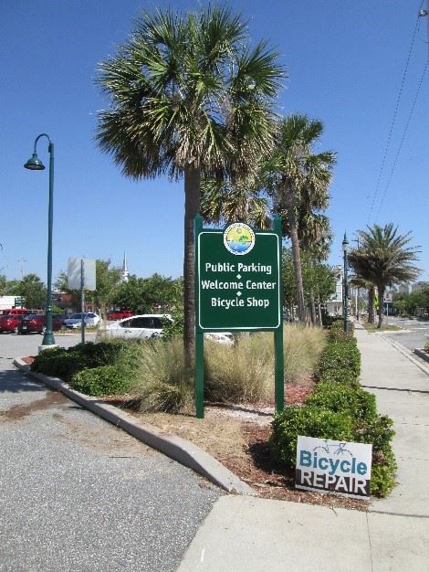 Photo of a large green sign in front of a parking lot, which reads: Public Parking - Welcome Center - Bicycle Shop